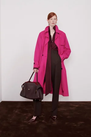 Model Laura Rudd wears a vibrant magenta trench coat and dark trousers while holding a large brown leather bag for Ferragamo.