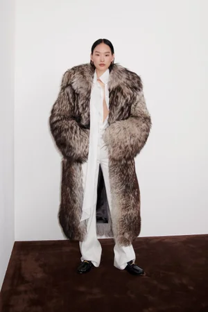Model wearing a voluminous fur coat over a white shirt and trousers against a plain studio background.