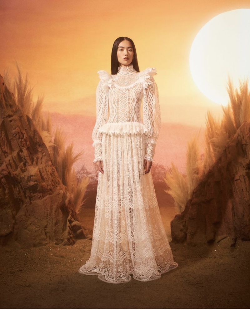 Model standing in a long white lace dress against a desert background with a glowing sun at sunset for Rodarte lookbook