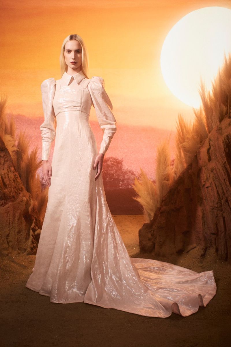 Woman in a shimmering, long-sleeved white gown posing in an outdoor setting with rocks, foliage, and a bright sun-like light in the background.