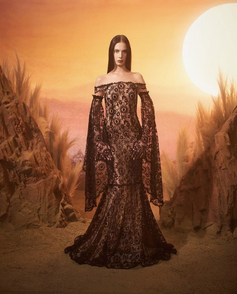 Brunette woman poses in black lace gown with off-the-shoulder neckline and long sleeves against desert landscape backdrop with large moon.