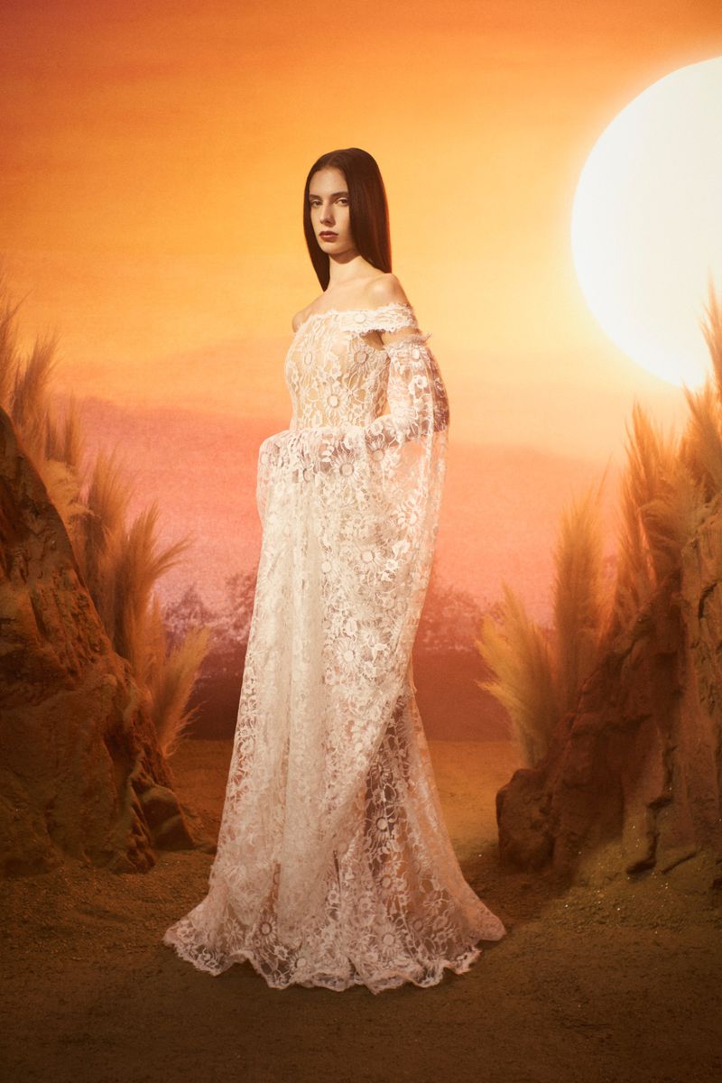 A woman in a lace off-the-shoulder gown poses in a surreal outdoor setting with a sunset and grassy knolls.