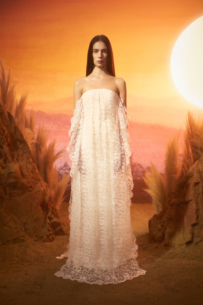 Elegant woman in a white lace dress standing in a scenic landscape with a soft, golden sunset and sparse vegetation.