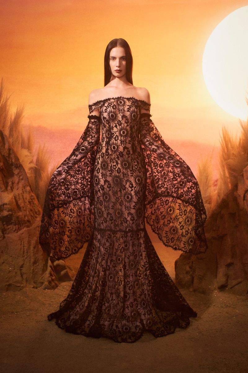 A brunette woman poses in a full-length, off-the-shoulder, black lace gown with sleeves, set against a warm-toned, surreal desert background.