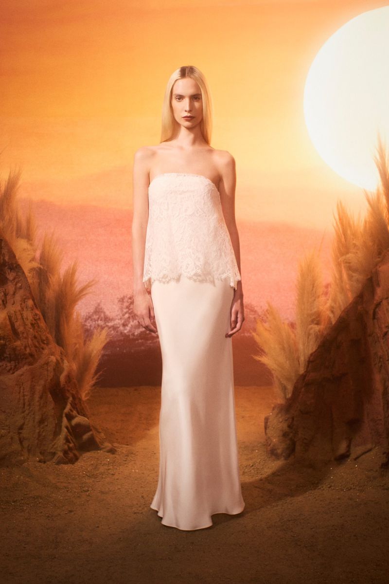 A fair-skinned woman in a strapless white gown stands in a desert landscape under a warm, stylized sun, fashion editorial.