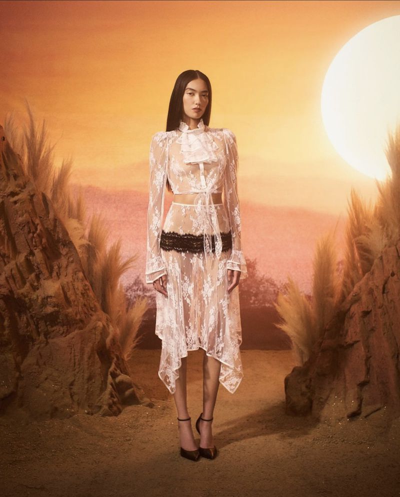A woman models a white lace dress against a sunset background, showcasing fashion with an ethereal and romantic aesthetic in a desert-like setting.
