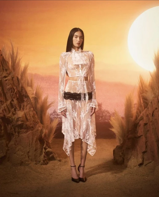 A woman models a white lace dress against a sunset background, showcasing fashion with an ethereal and romantic aesthetic in a desert-like setting.