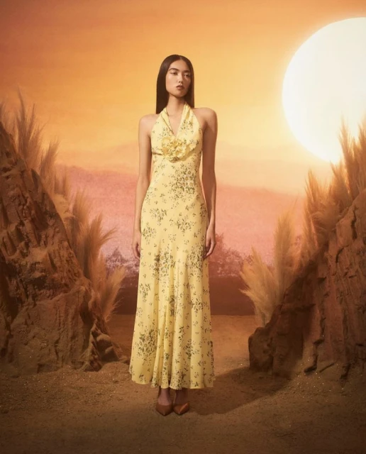 A slender woman poses in a yellow halter dress featuring a floral pattern against a warm-toned background with dry grasses and rock formations.