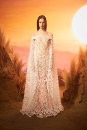 Model wearing an off-the-shoulder white lace gown standing against a sunset sky and rocky terrain backdrop.