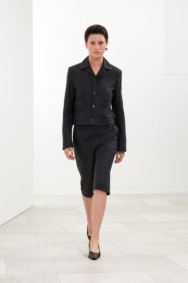 A slender woman walks toward the camera in a stark white indoor setting, wearing a dark jacket, capri pants, and black shoes.
