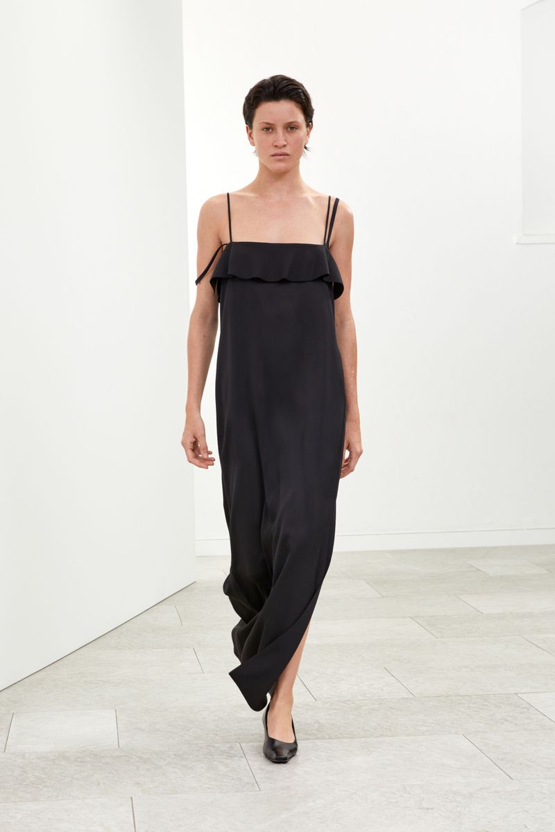 Fashion shot of a slender woman in a black dress walking against a minimalist white background, showcasing modern elegance and style.