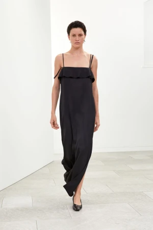 Fashion shot of a slender woman in a black dress walking against a minimalist white background, showcasing modern elegance and style.