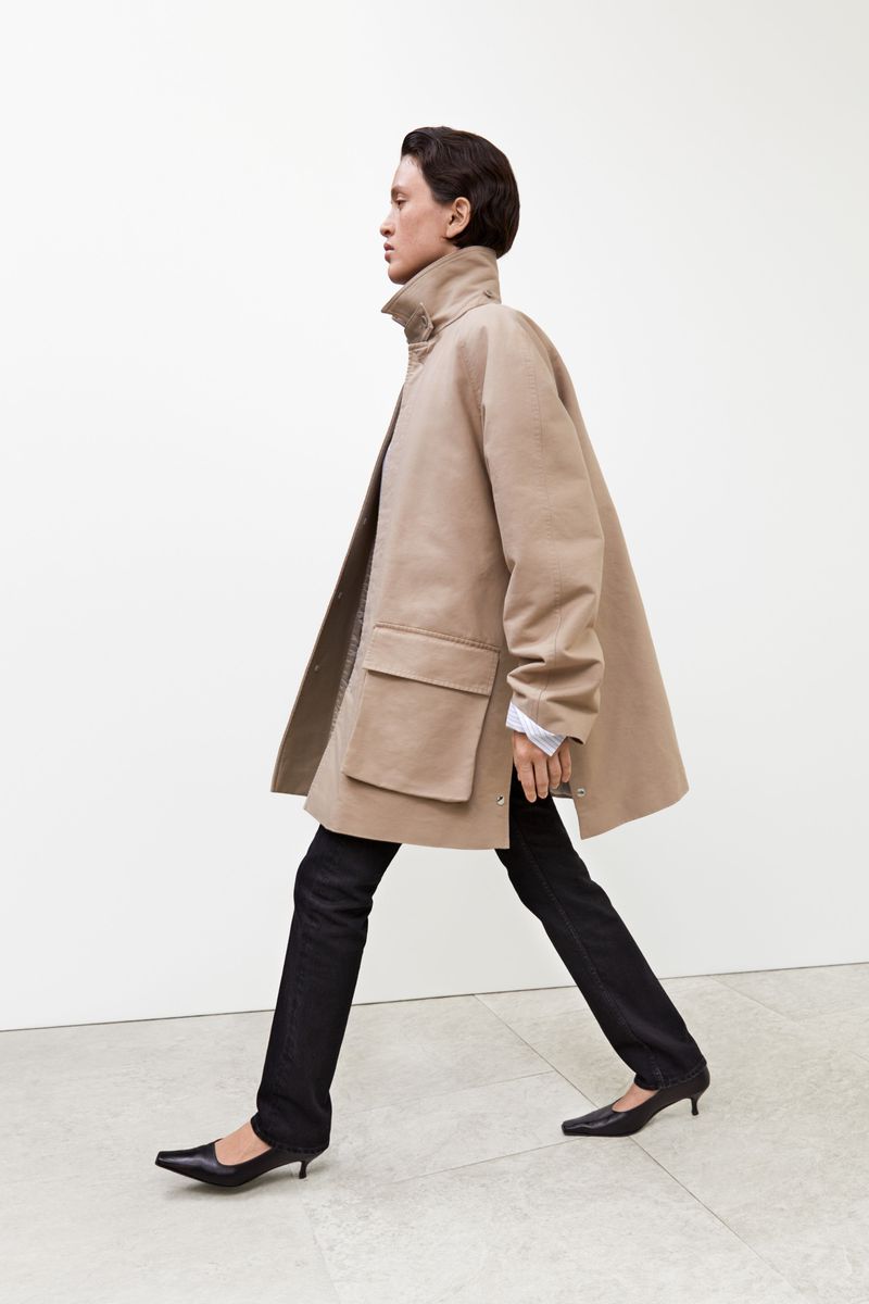 Fashion editorial shot of a woman walking, wearing a tan coat, dark pants and black heeled shoes, against a neutral studio backdrop.