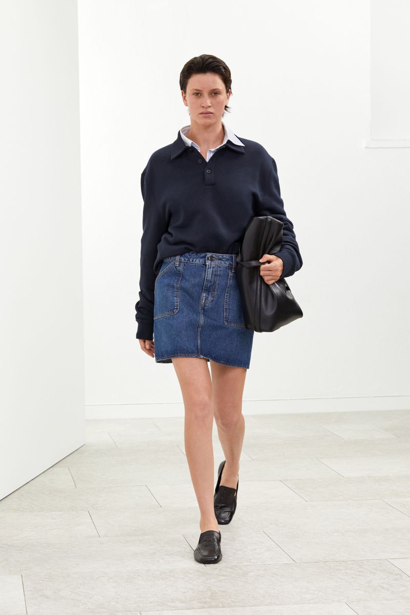 A female model walks in a white interior, wearing a navy blue polo shirt, denim skirt, black loafers and carrying a black leather bag.