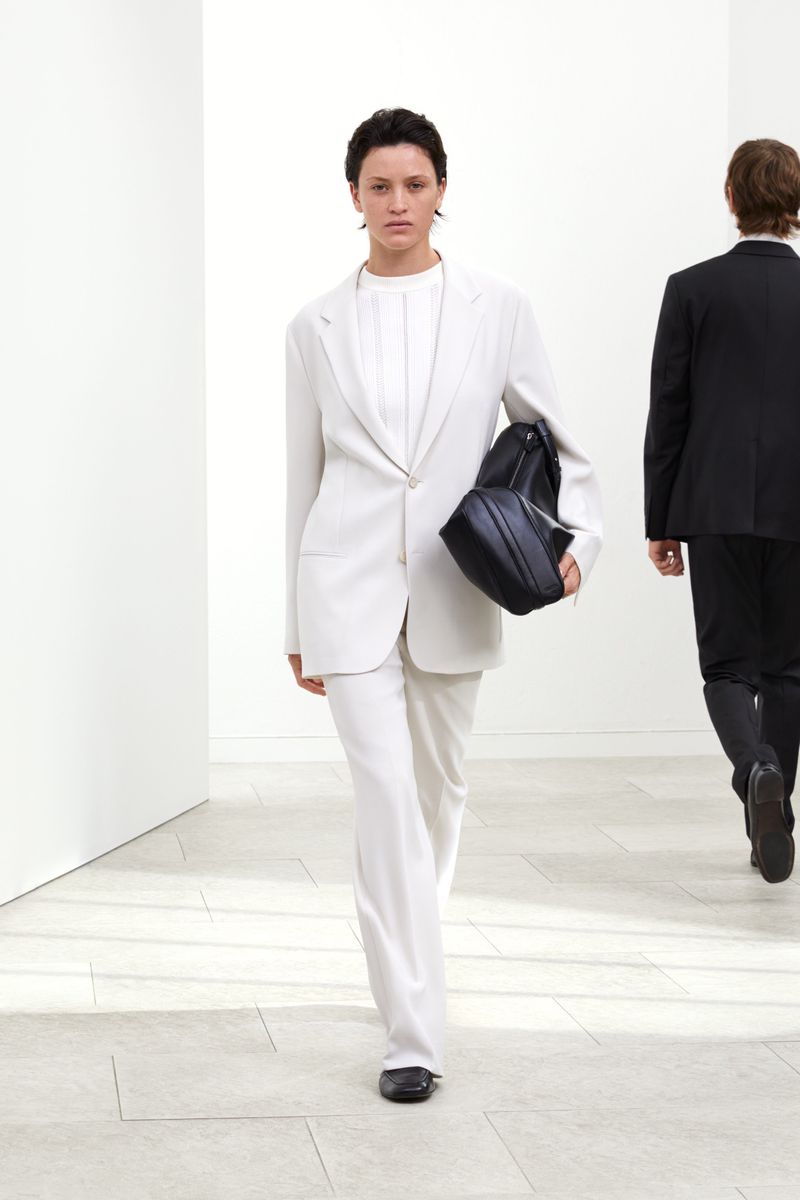 A woman in an all-white suit and a black bag walks toward the camera in a bright corridor with another person in the background.