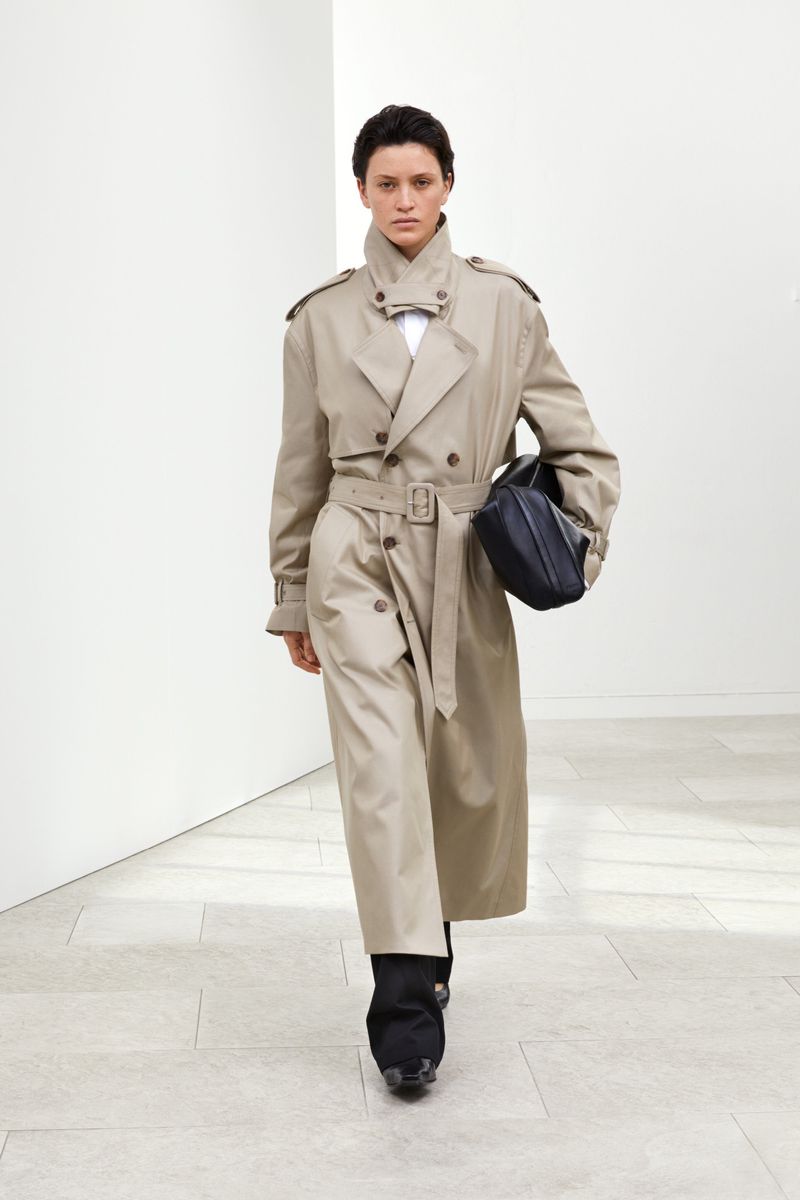 A fashion-forward woman in a trench coat strides confidently in a minimalist white studio, holding a black bag, showcasing modern elegance.