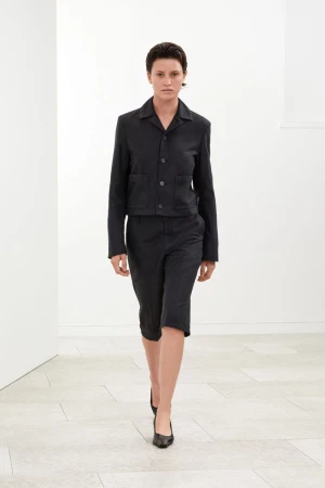 Model Puck Schrover walking in a black cropped jacket, matching knee-length shorts, and black pointed heels in a white studio room.