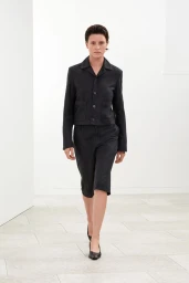 Model Puck Schrover walking in a black cropped jacket, matching knee-length shorts, and black pointed heels in a white studio room.