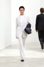 Model walking forward in an off-white tailored trouser suit carrying a black bag under arm in a bright minimalist interior.