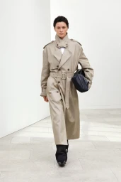 Model in a long belted beige trench coat walking forward against a white architectural background