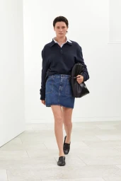 Model walking forward wearing a navy polo sweater, denim skirt, and black loafers while holding a black leather bag