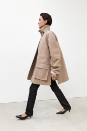 Model walking in profile wearing a structured beige oversized coat, black trousers, and pointed kitten heels against a white wall.