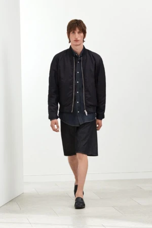 Male model walking in a black bomber jacket, dark button-down shirt, black shorts, and leather loafers against a white backdrop.