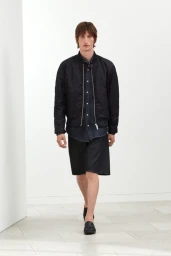 Male model walking in a black bomber jacket, dark button-down shirt, black shorts, and leather loafers against a white backdrop.