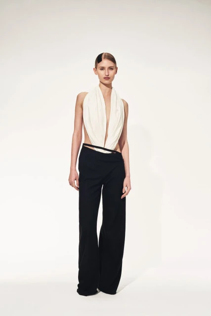 Full-length shot of a model posing in a cream halter top and black pants against a stark white background in a fashion editorial style.