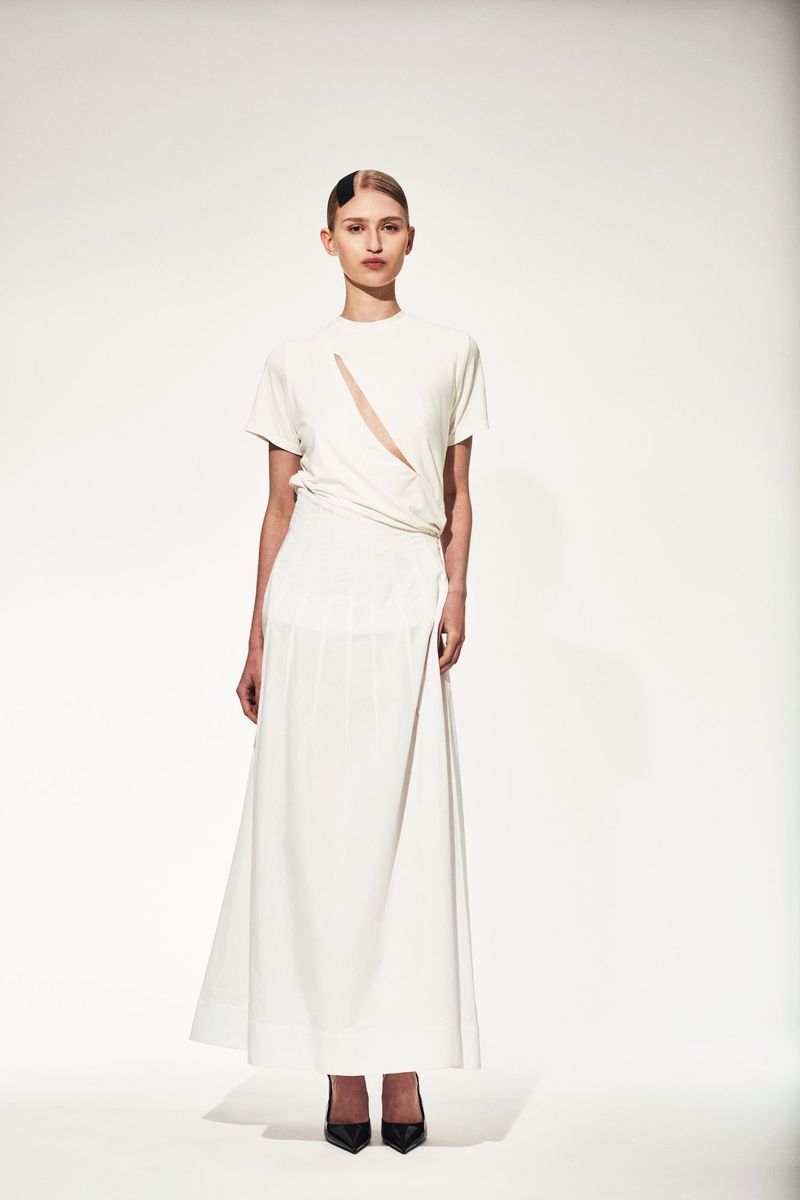 Full-length portrait of a slender fashion model in an off-white short-sleeve top with asymmetrical neckline, and a matching maxi skirt.