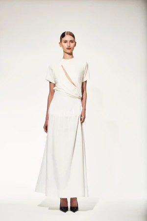 Full-length portrait of a slender fashion model in an off-white short-sleeve top with asymmetrical neckline, and a matching maxi skirt.