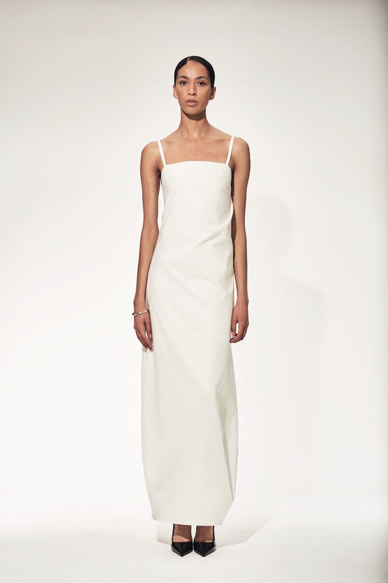 Elegant adult woman in a full-length white dress posing in a light studio. Fashion shot with neutral tones and clean background.