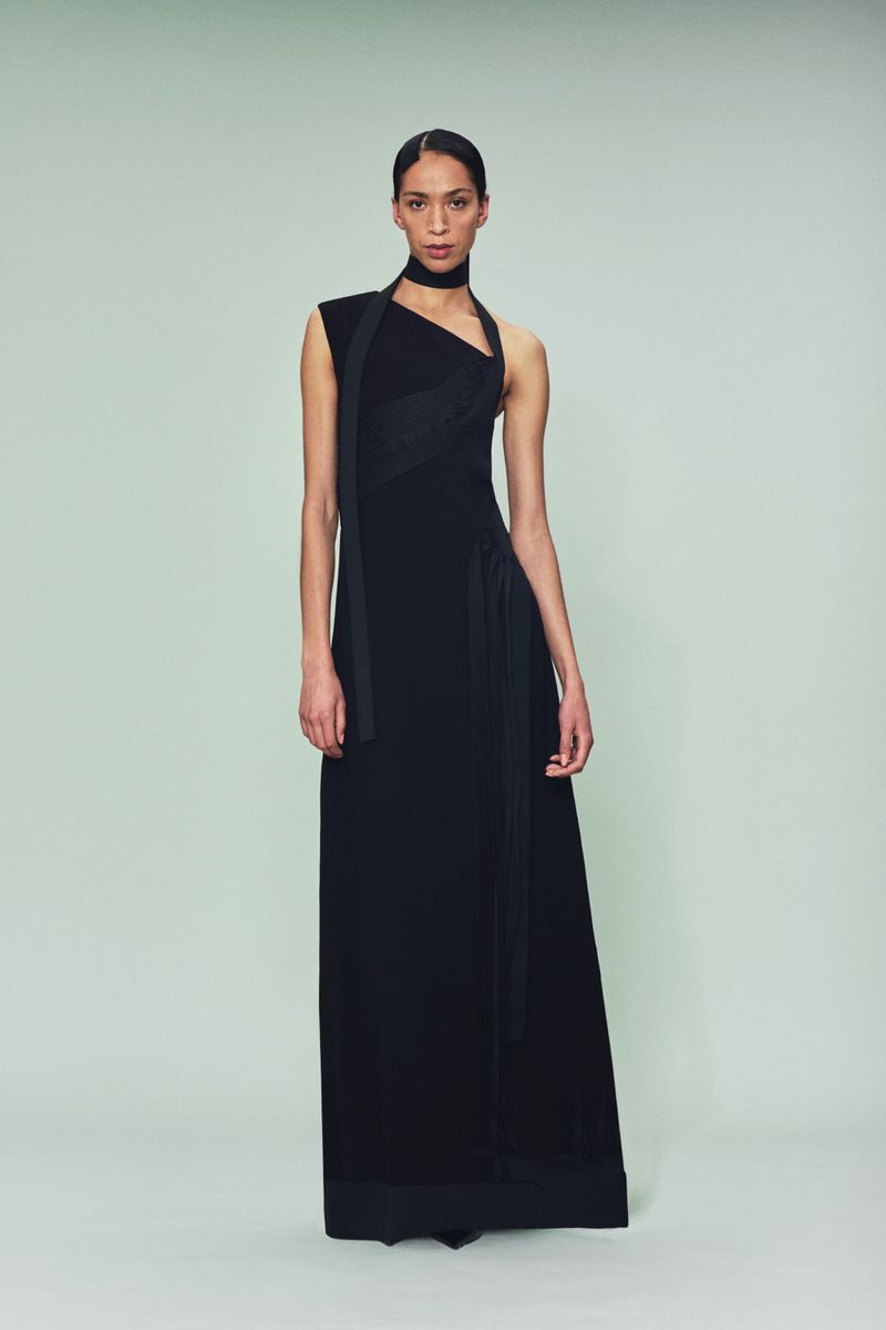 A woman poses in a full-length black gown with a matching choker against a neutral background, showcasing a sleek and elegant design.