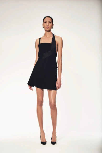 A female fashion model poses in a black mini dress and high heel shoes against a white backdrop in a studio setting.