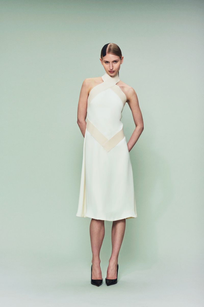 Full-length studio shot of a young, fair-skinned woman in a cream-colored dress and black heels, posed against a pastel green backdrop.