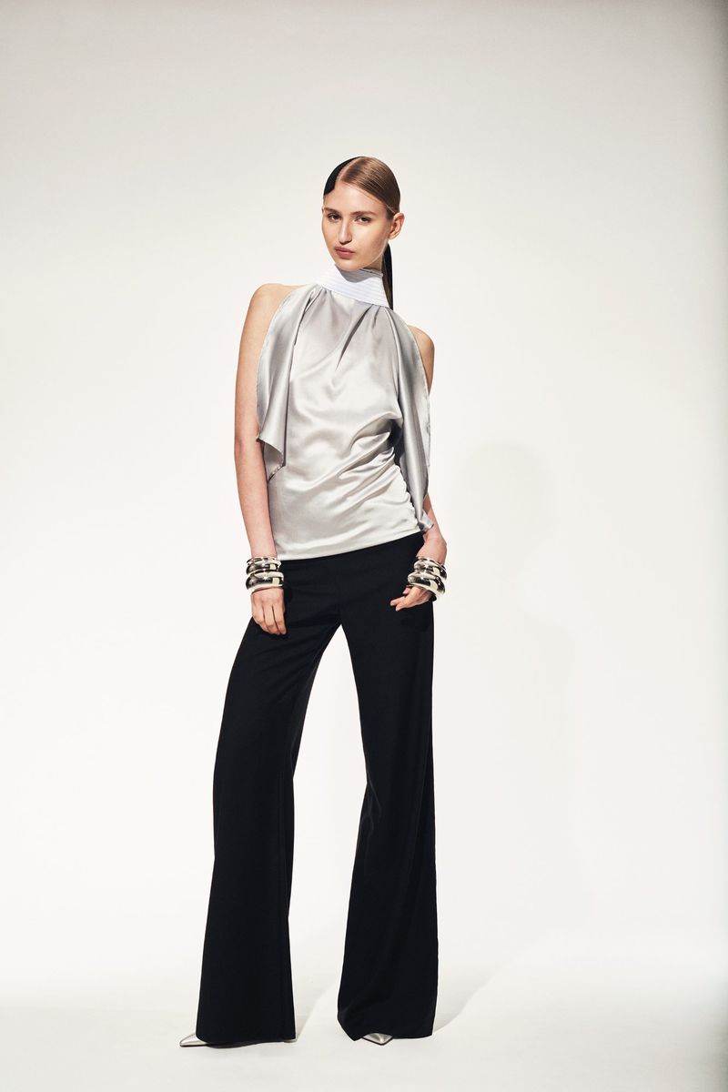 A slender woman in a silver halter top and black wide-leg pants poses in a fashion studio with a clean, white backdrop, accessorized with bracelets.