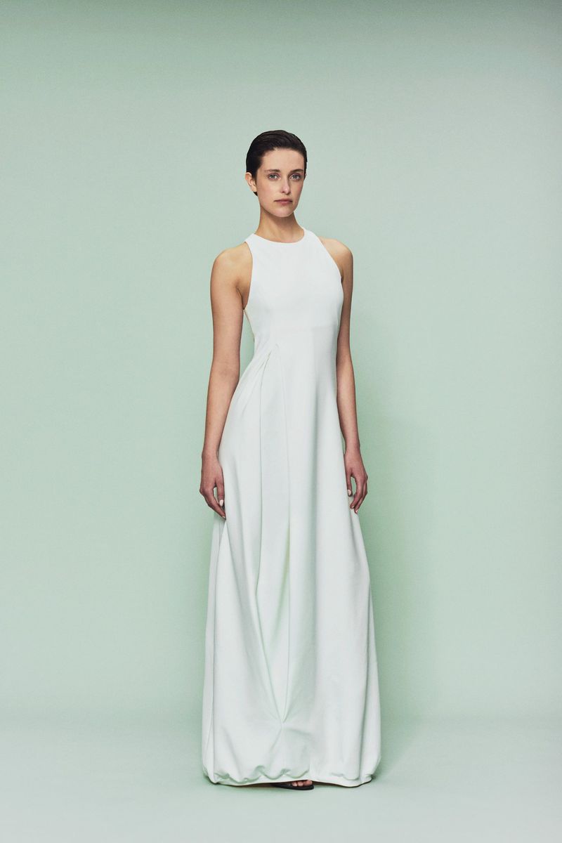 Full-length portrait of a slender female fashion model in a sleeveless, light-mint green gown, posing against a light green backdrop.