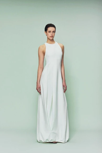 Full-length portrait of a slender female fashion model in a sleeveless, light-mint green gown, posing against a light green backdrop.