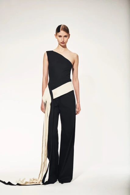 Elegant adult female fashion model poses in black one-shoulder jumpsuit with cream sash and fringe against a clean white backdrop.