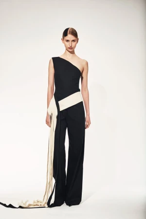 Elegant adult female fashion model poses in black one-shoulder jumpsuit with cream sash and fringe against a clean white backdrop.