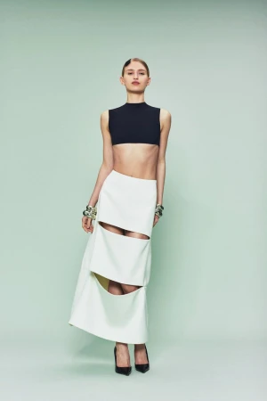 Model wearing a black sleeveless crop top and a white cutout skirt with black pointed heels against a pale green backdrop.