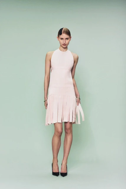 Full-length portrait of a woman in a light pink dress and black heels, against a light green studio backdrop. Fashion editorial shot.