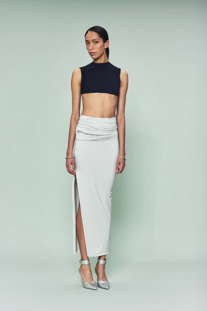 Woman poses in a black crop top and light gray skirt with a thigh-high slit, standing against a solid light green backdrop.