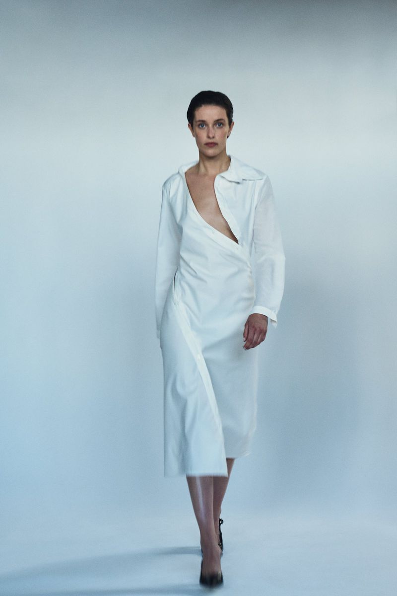 A woman in a white dress walks toward the camera. She has short hair and is wearing high heel shoes. The background is a light grey.