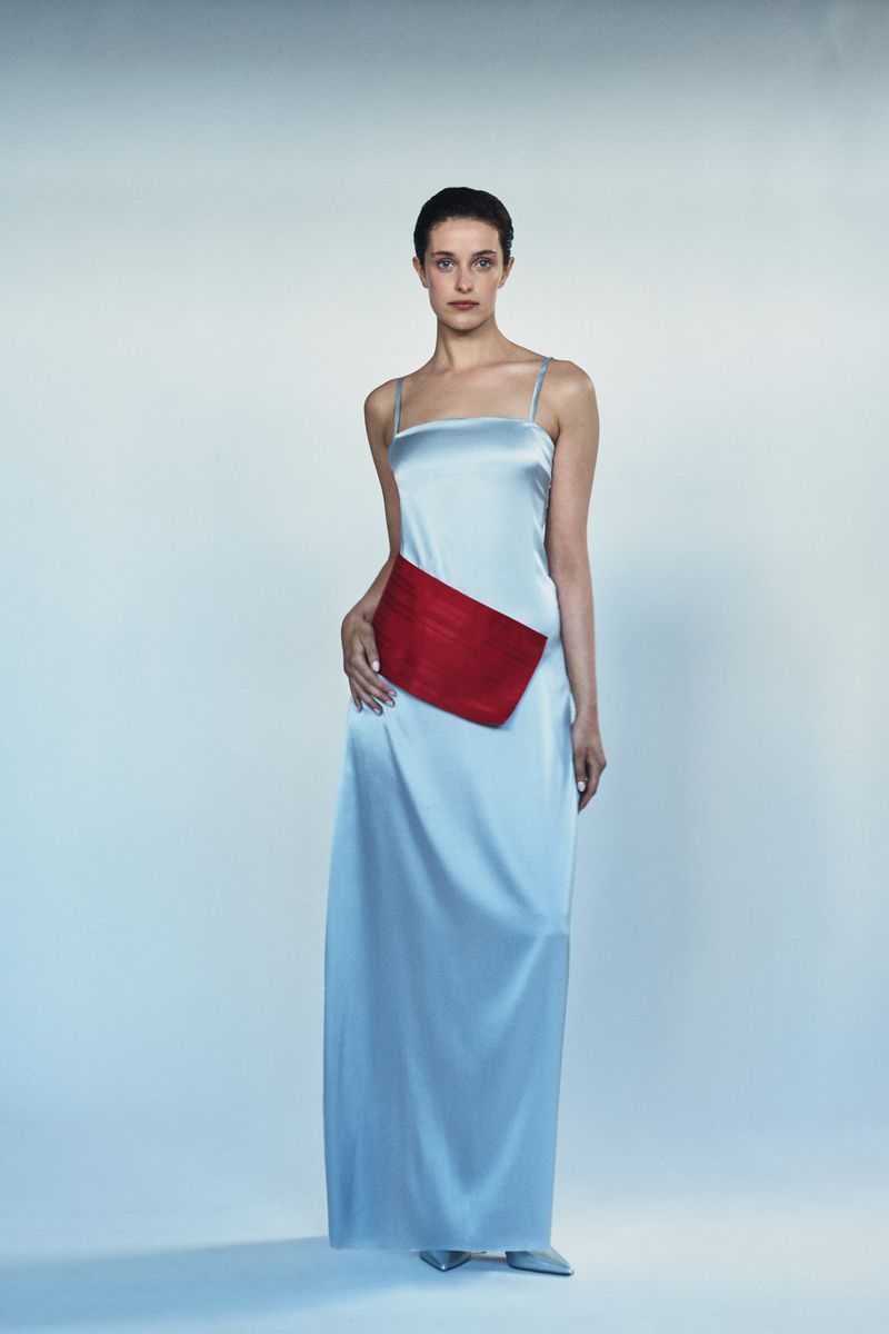 A woman stands in a full-length light blue satin dress with a red sash. She is posed against a solid pale blue background.