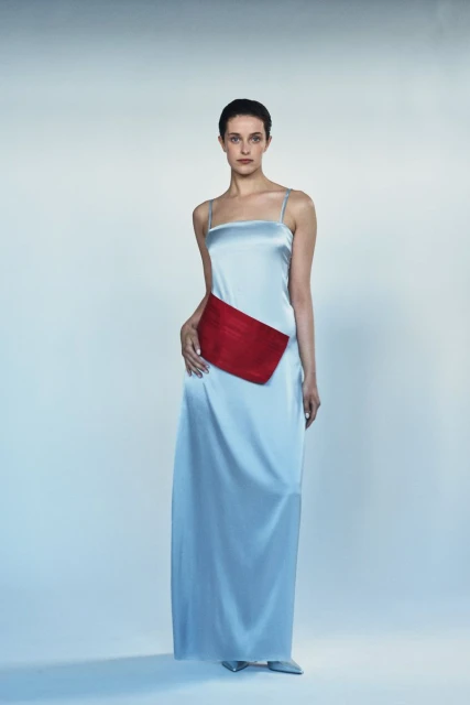 A woman stands in a full-length light blue satin dress with a red sash. She is posed against a solid pale blue background.