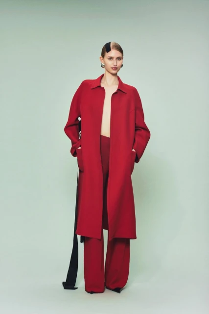 Fashionable adult woman poses in a red coat and matching pants against a pale green backdrop, wearing a black hair clip and earrings.