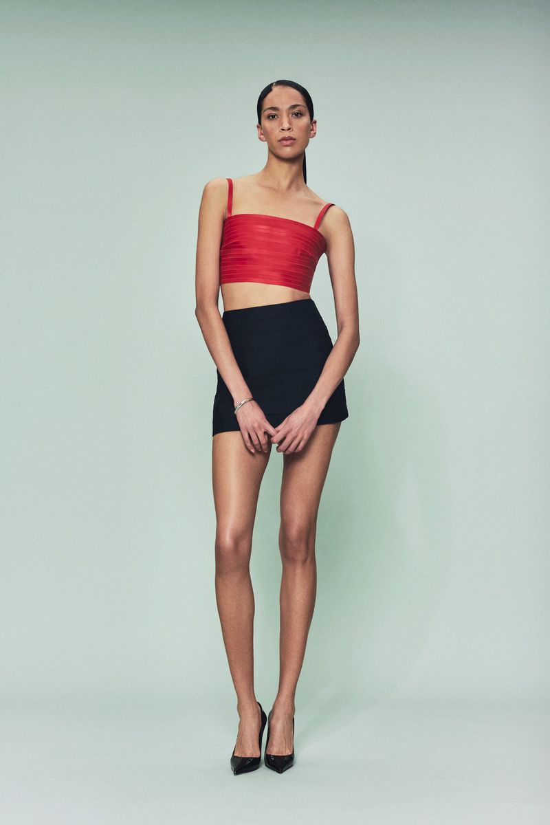 Fashion model poses in a red top and black skirt against a solid green background in a studio shot, showcasing modern apparel.