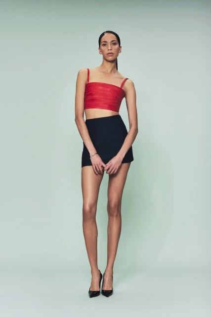 Fashion model poses in a red top and black skirt against a solid green background in a studio shot, showcasing modern apparel.