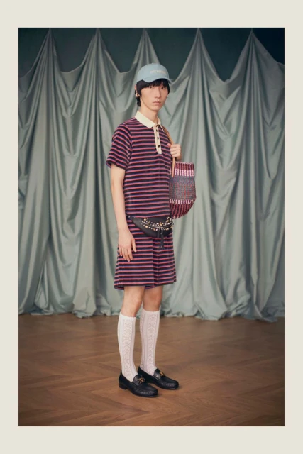 A person stands wearing a striped collared dress, belt bag, cap, knee-high socks and loafers, carrying a multicolored woven tote bag.